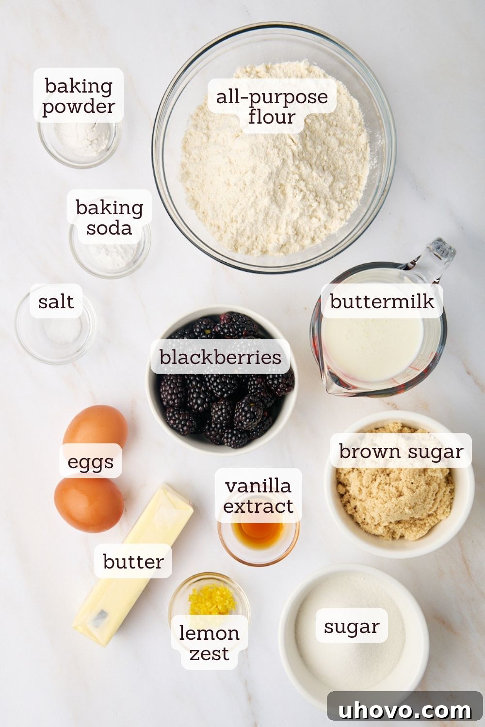 An overhead view of various baking ingredients for blackberry muffins laid out on a surface, including flour, sugar, eggs, buttermilk, and fresh blackberries.