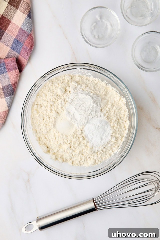 An overhead view of flour, baking powder, baking soda, and salt measured in a clear glass mixing bowl, ready to be whisked together for the muffin batter.