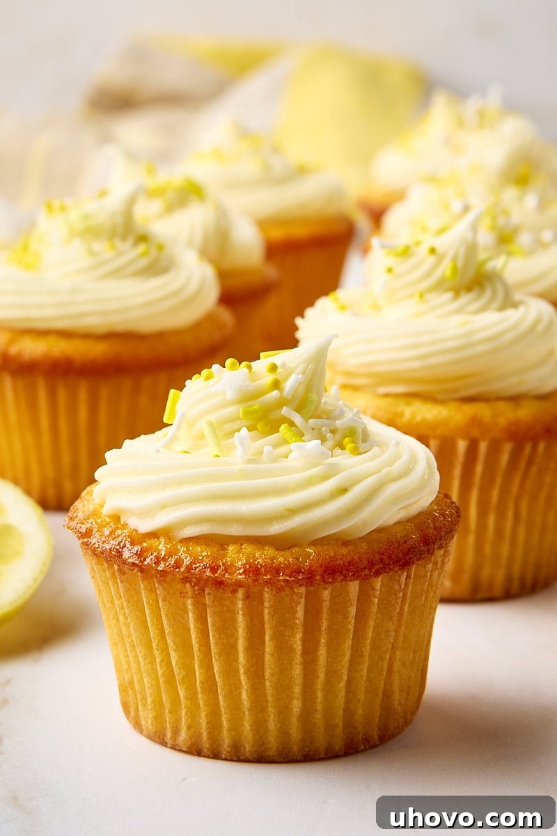 A beautifully frosted lemon cupcake, bright yellow, sitting on a white surface, with more blurry cupcakes in the background, hinting at a fresh batch ready to be enjoyed.