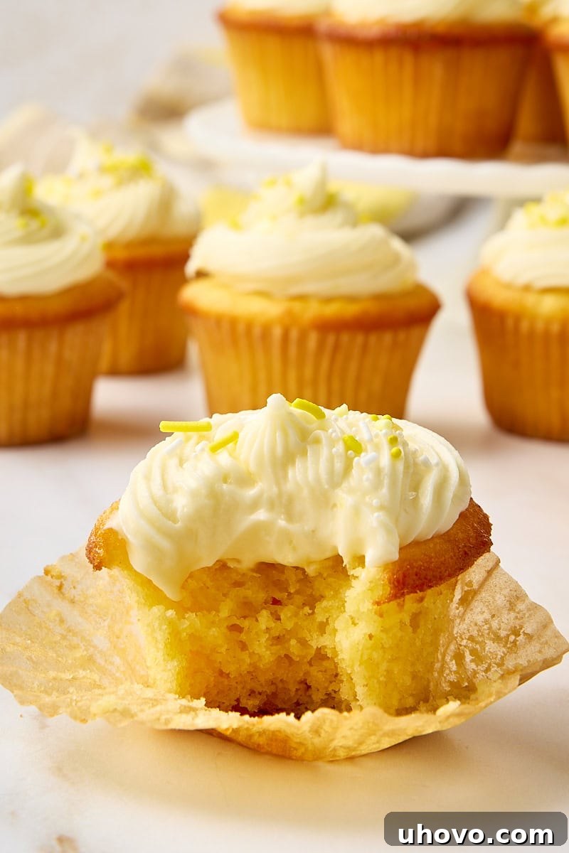 A close-up of a lemon cupcake with a bite taken out, revealing its fluffy interior, with several more frosted cupcakes blurred in the background, enticing viewers to take a bite.