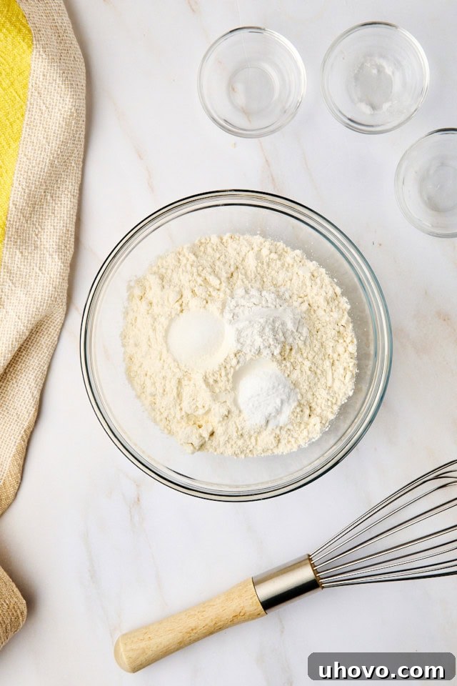 Overhead view of a glass mixing bowl containing pre-measured flour, baking powder, baking soda, and salt, ready to be whisked together.