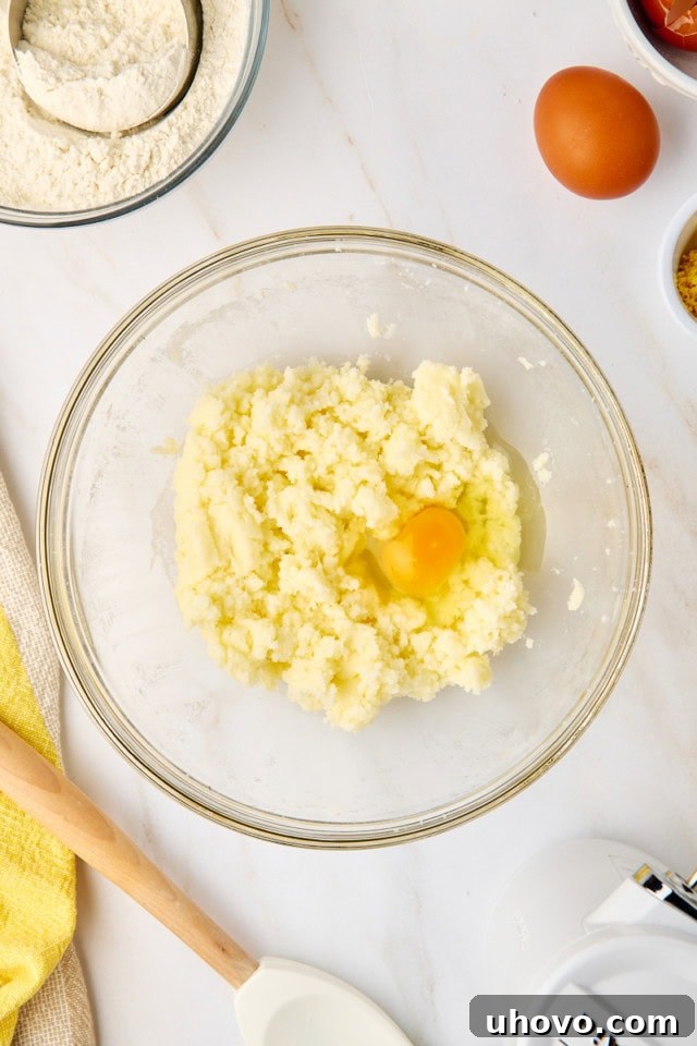 Close-up, overhead view of a cracked egg being added to a creamy mixture of butter and granulated sugar in a stand mixer bowl.