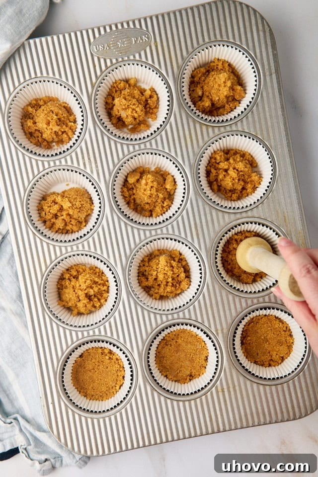 An overhead shot showing the graham cracker crust mixture being meticulously pressed into the bottoms of lined muffin cups, forming the sturdy base for the cheesecakes.