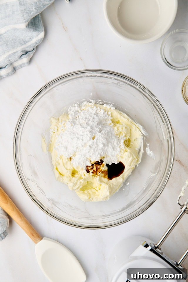 An overhead view of confectioners' sugar, vanilla bean paste, and a touch of lemon juice being added to beautifully beaten cream cheese in a clear glass mixing bowl, signaling the start of the filling.