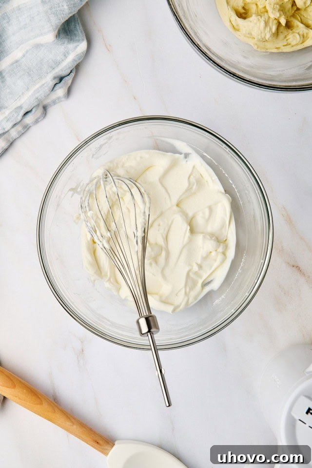 An overhead shot showcasing perfectly whipped heavy cream with stiff peaks forming in a separate glass mixing bowl, ready to be incorporated into the cheesecake filling.