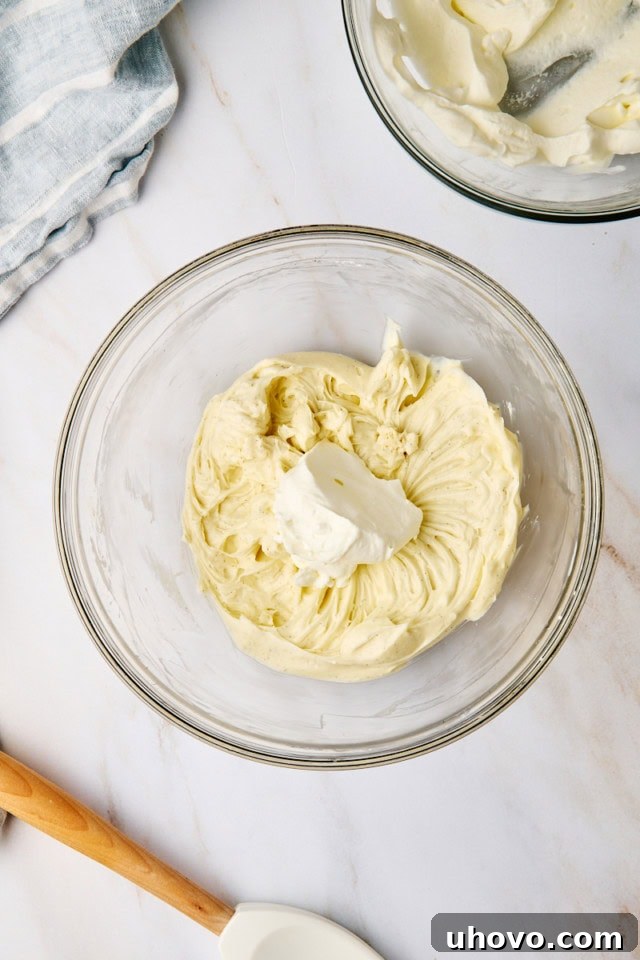 An overhead view capturing a portion of the whipped cream being gently folded into the cream cheese mixture in a glass mixing bowl, ensuring a light and airy texture.