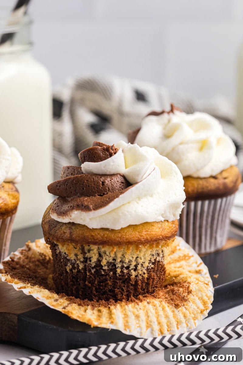 An unwrapped marble cupcake on a black surface, highlighting its distinct chocolate and vanilla swirl, with more cupcakes in the blurred background. It promises a delightful blend of flavors.
