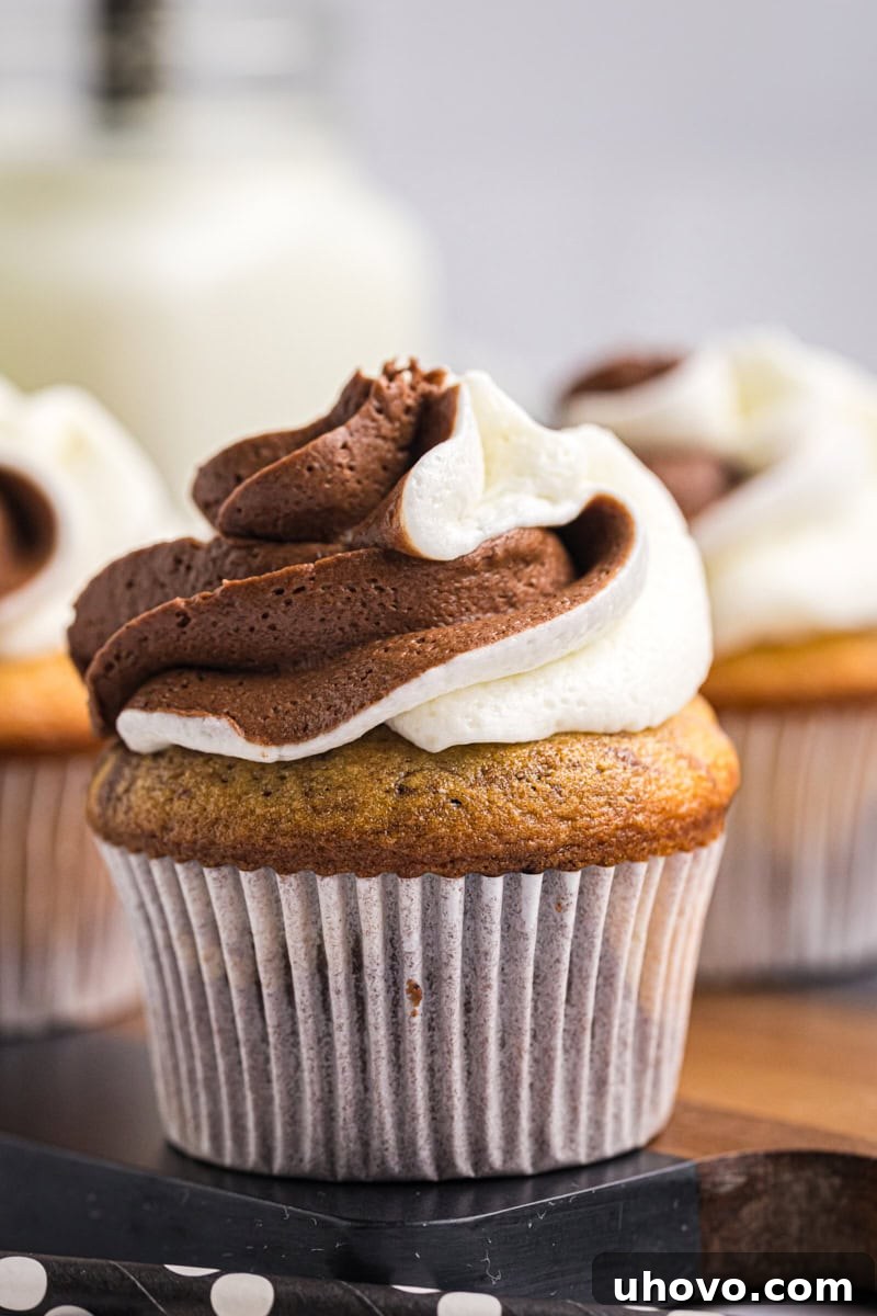 A close-up shot of a single marble cupcake with its two-tone frosting, resting elegantly on a wooden surface, with more cupcakes partially visible and softly blurred in the background. The focus is on the intricate details of the frosting and cake.