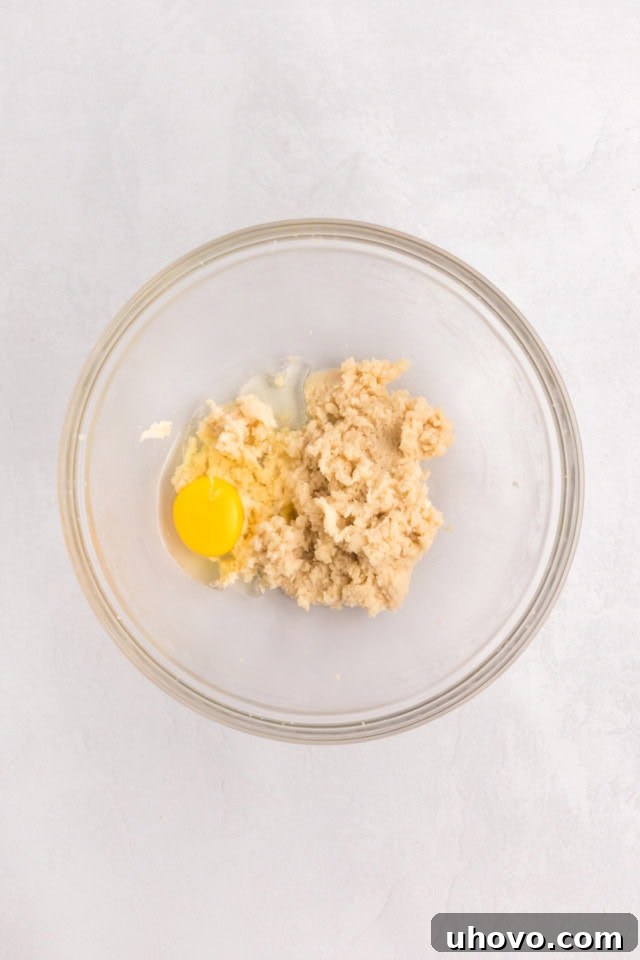 An overhead view of a large glass mixing bowl containing creamed butter and sugars, with an egg being added to the mixture, ready for beating. This is the initial step for the cupcake batter.