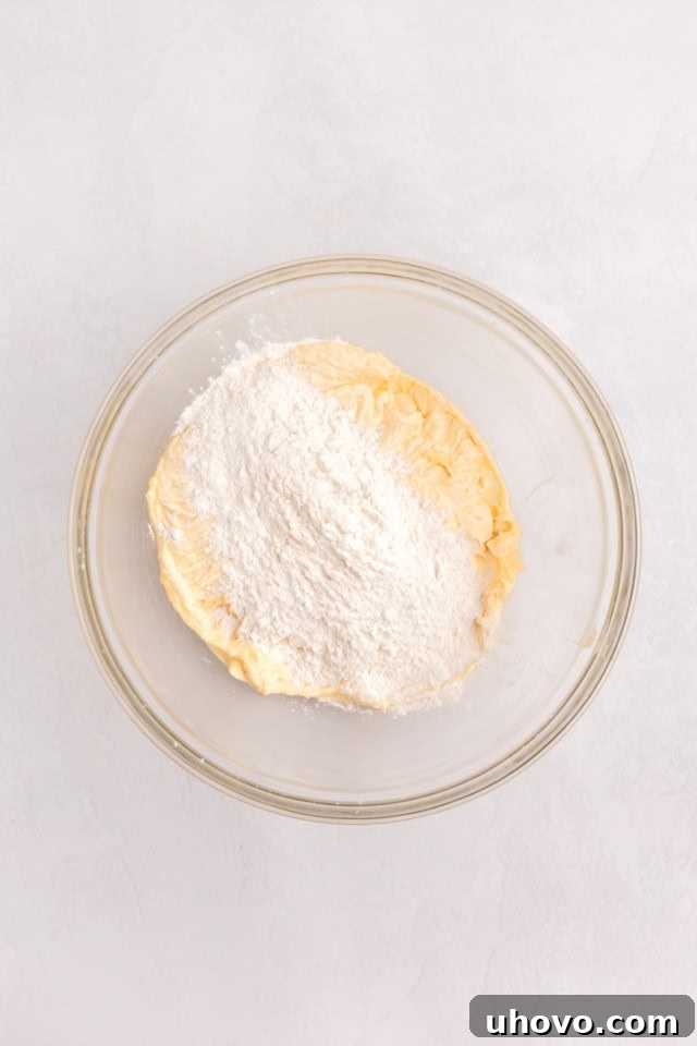 An overhead view of dry ingredients, including flour, baking powder, baking soda, and salt, being added in portions to the wet ingredients in a glass mixing bowl, forming the cupcake batter.