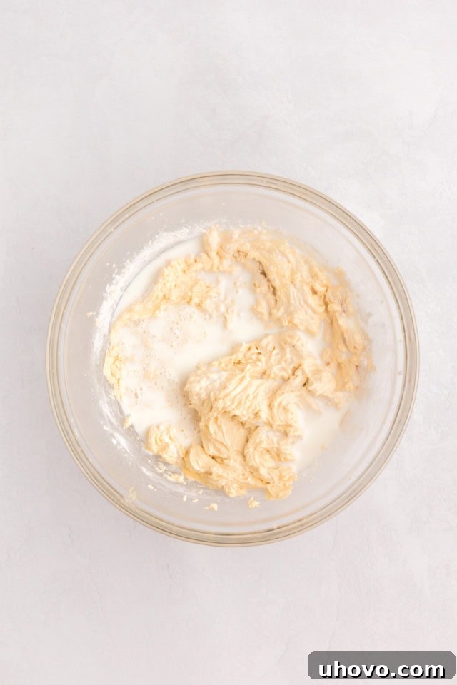 An overhead view of buttermilk being poured into a glass mixing bowl filled with cupcake batter, demonstrating the alternation of wet and dry ingredients.