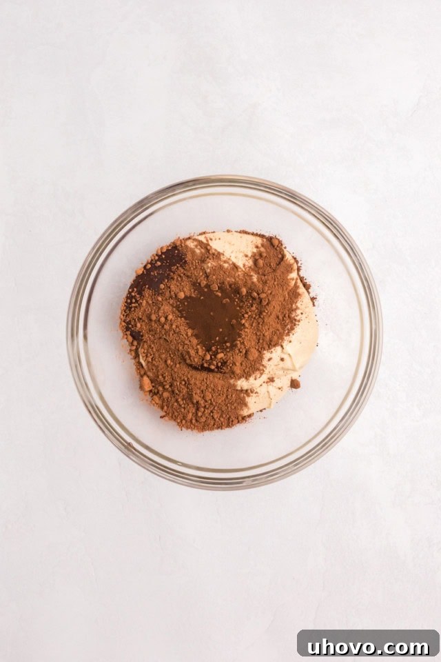 An overhead view of cocoa powder, melted chocolate, and optional espresso powder being added to a portion of vanilla cupcake batter in a glass mixing bowl, ready to create the chocolate swirl.