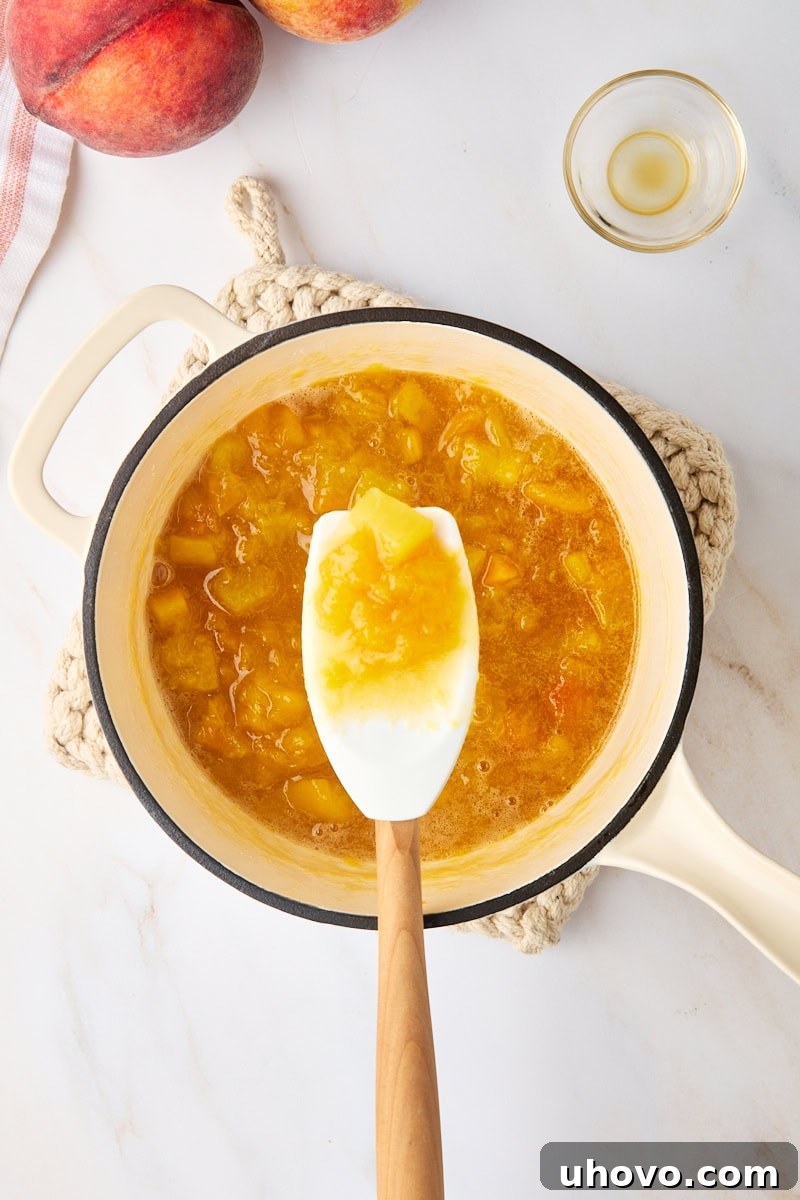 Overhead shot of a spoonful of glistening peach compote, held above the remaining compote in a saucepan, showing its perfect consistency.