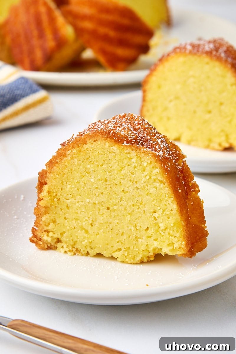 A perfectly baked slice of cornmeal pound cake, elegantly topped with a dusting of powdered sugar, presented on a white plate with more cake in the soft-focus background.