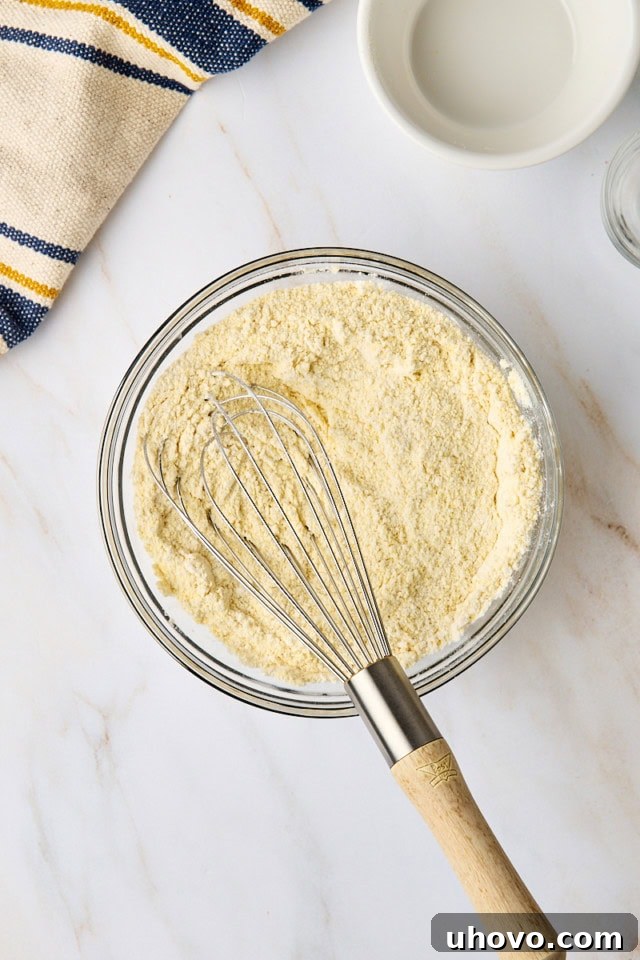 An overhead view of flour, cornmeal, baking powder, and salt meticulously whisked together in a clear glass mixing bowl, ensuring even distribution.