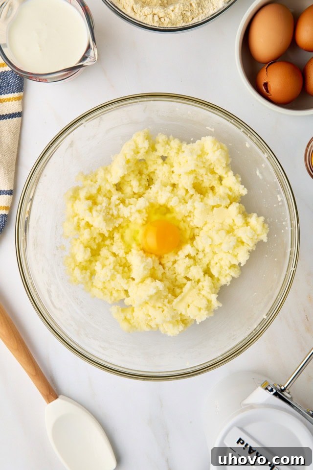 An overhead view of a single large egg being added to the light and fluffy creamed butter and sugar mixture in a glass mixing bowl, a key step in building emulsion.