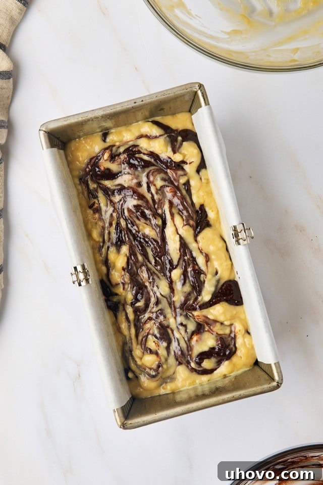 Overhead view of marbled chocolate banana bread batter in a loaf pan, showing alternating spoonfuls of light and dark batter before swirling, ready for the oven.