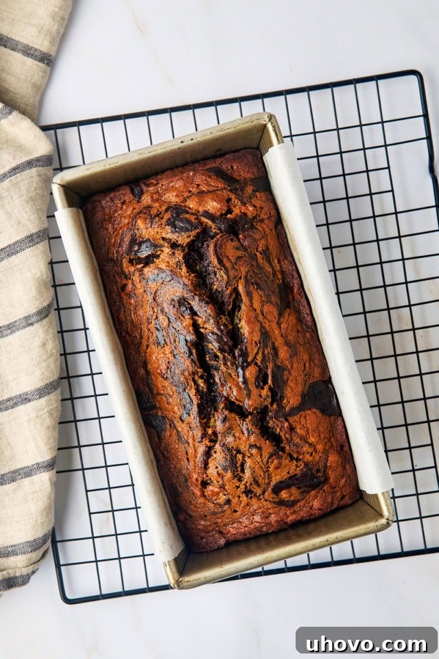 Overhead view of a freshly baked loaf of marbled chocolate banana bread in a loaf pan, cooling on a wire rack, showcasing a beautiful golden crust and visible swirls.