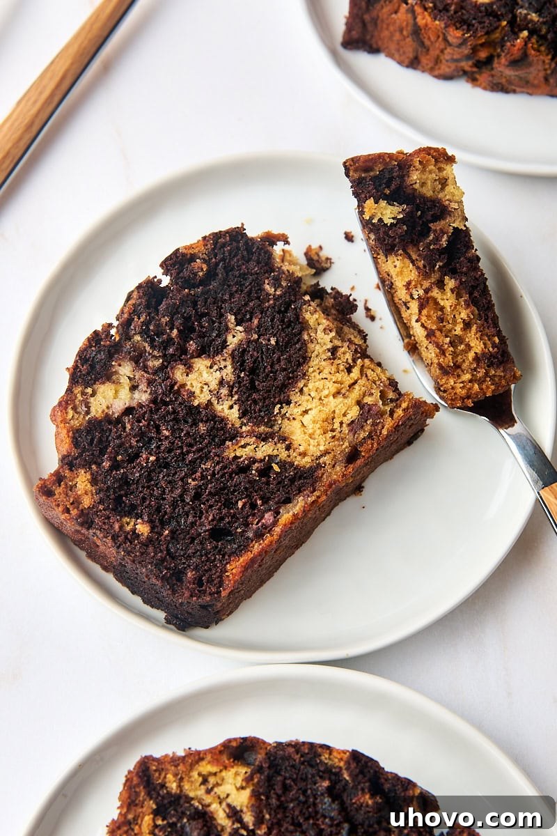 A close-up of a slice of marbled chocolate banana bread on a white plate, with a fork holding a bite, emphasizing its moist interior and perfect swirl.