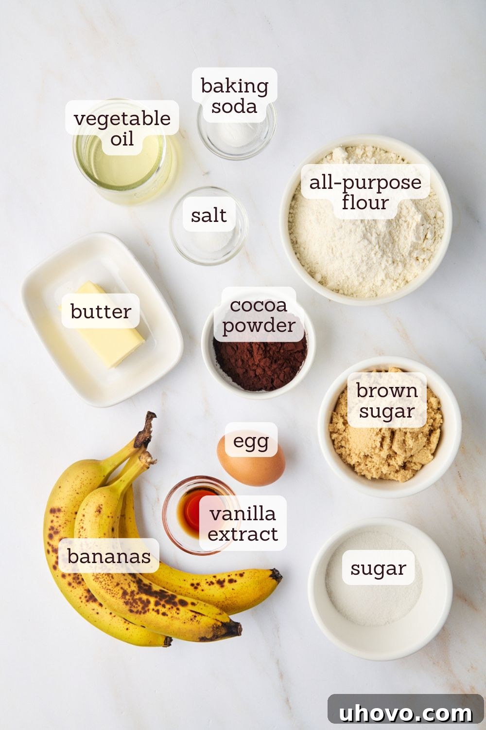 Overhead view of all the simple ingredients laid out for making marbled chocolate banana bread: bananas, flour, sugar, butter, oil, cocoa powder, and eggs.