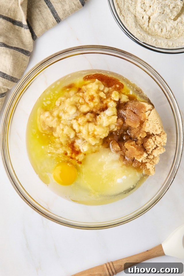 Wet ingredients for marbled chocolate banana bread, including mashed bananas, sugars, egg, melted butter, oil, and vanilla extract, are shown stirred together in a large glass mixing bowl.