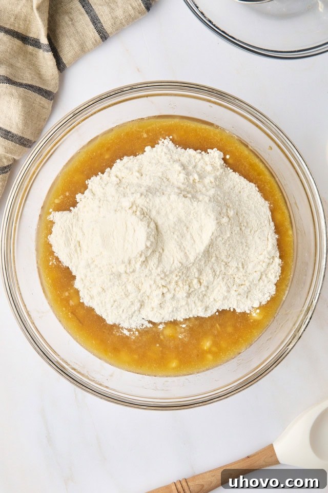 Overhead view of dry ingredients for banana bread being poured into the wet ingredients in a glass mixing bowl, ready to be gently stirred together.
