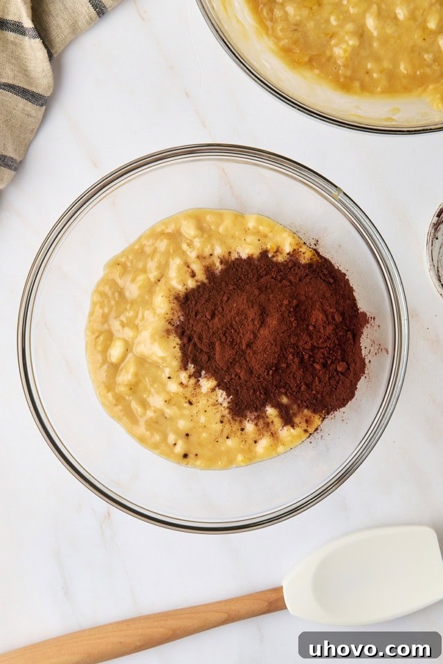 Overhead view of unsweetened cocoa powder being added to half of the banana bread batter in a glass mixing bowl, preparing it for the chocolate swirl.