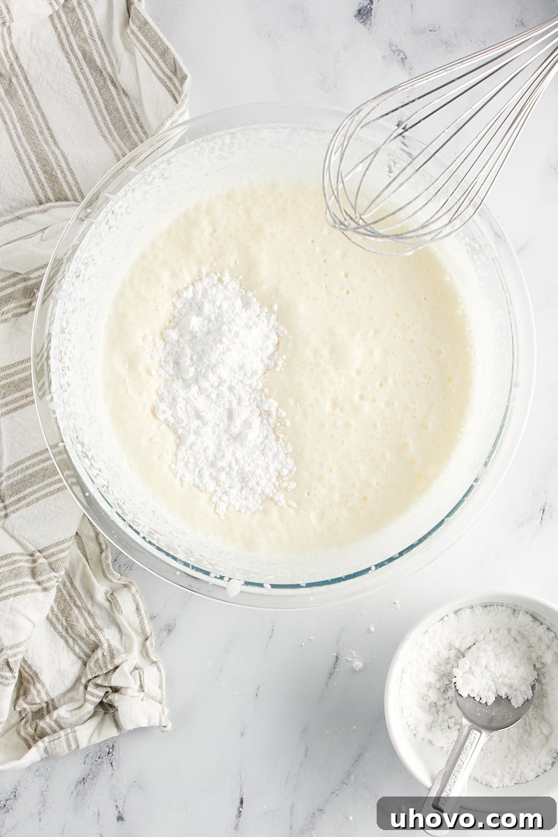 overhead view of confectioners' sugar added to partially whipped cream in a glass mixing bowl