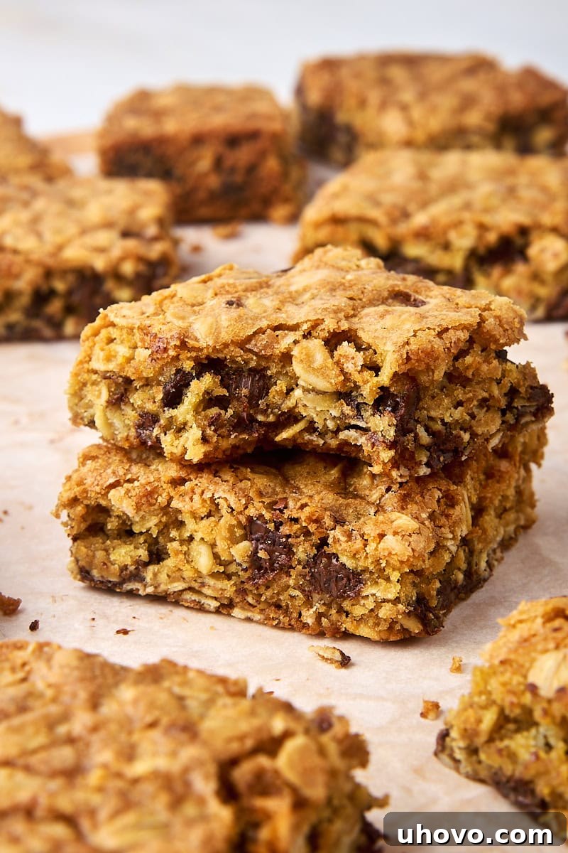 A close-up of two stacked oatmeal cookie bars, with a bite taken from the top one, revealing its soft, chewy interior filled with oats and chocolate. More bars are blurred in the background.