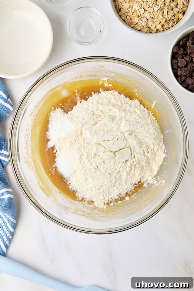 A glass mixing bowl with the wet ingredients, and a measuring cup with dry ingredients (flour, baking soda, salt) being added to the bowl.