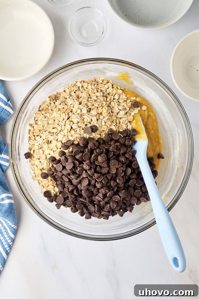 Old-fashioned rolled oats and chocolate chips being poured into the cookie batter in a glass mixing bowl.