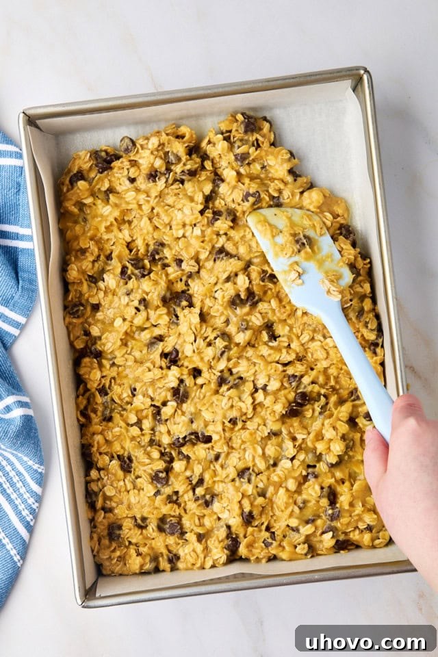 Overhead view of oatmeal cookie bar batter being carefully spread and pressed into a parchment-lined 9x13 inch baking pan.