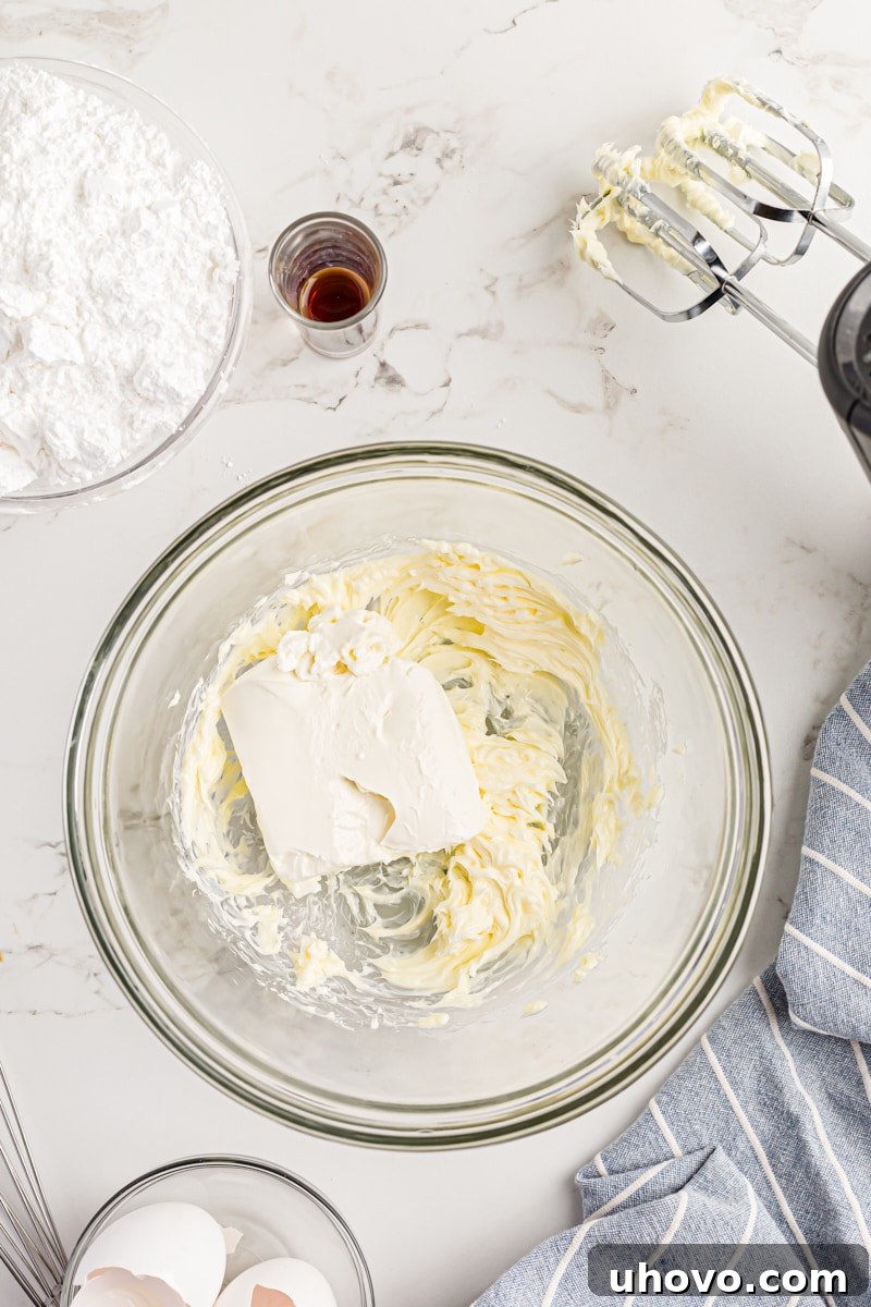 Overhead view of a block of softened cream cheese being added to whipped unsalted butter in a mixing bowl.