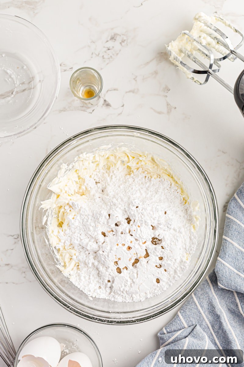 Overhead view of vanilla extract and sifted confectioners' sugar being added to the butter and cream cheese mixture.
