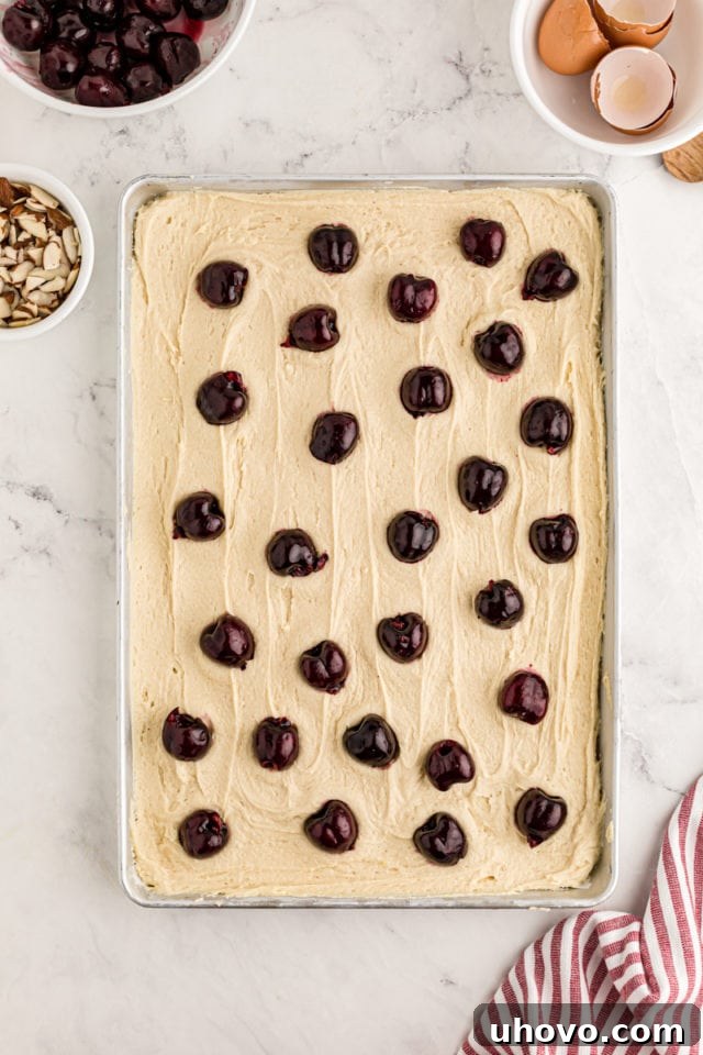 An overhead shot of the cake batter expertly spread evenly into the prepared sheet pan, demonstrating the smooth foundation laid before the addition of cherries and almonds.