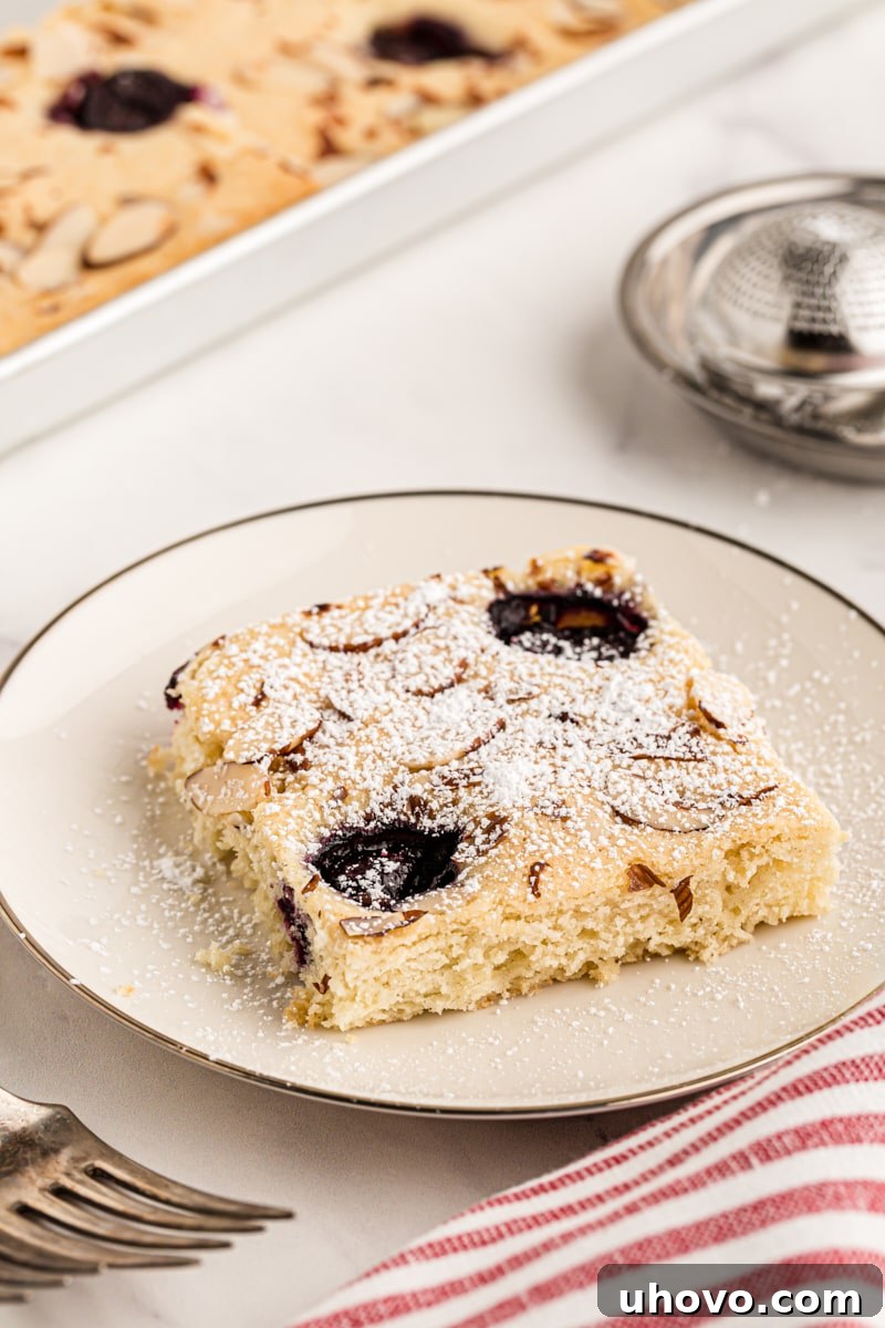 A perfectly sliced piece of cherry almond sheet cake on a white plate, generously dusted with powdered sugar, inviting indulgence.