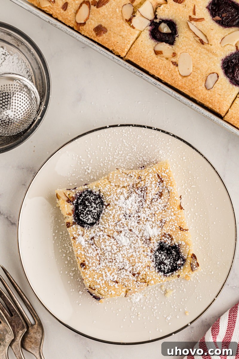 An inviting overhead view of a cherry almond sheet cake, partially served on a plate, with the remaining cake beautifully arranged in its sheet pan. Fresh cherries scattered nearby emphasize the cake's vibrant, fruity essence.