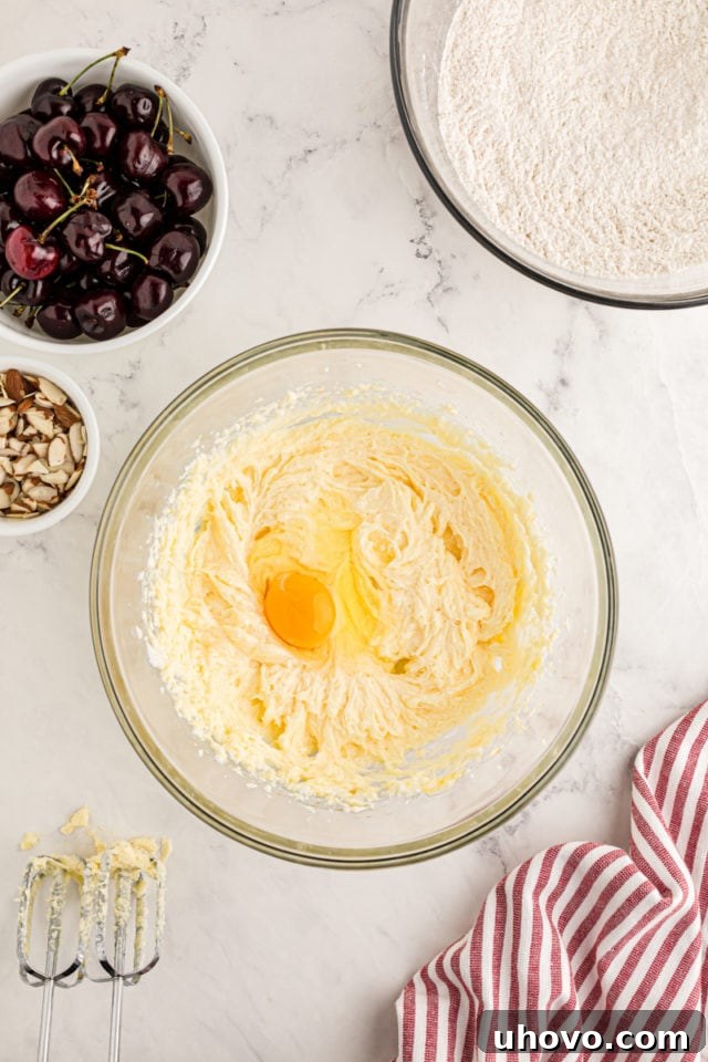 An egg being carefully cracked into a bowl already containing the creamy mixture of butter and sugar, illustrating the sequential addition of wet ingredients to build the cake batter's foundation.