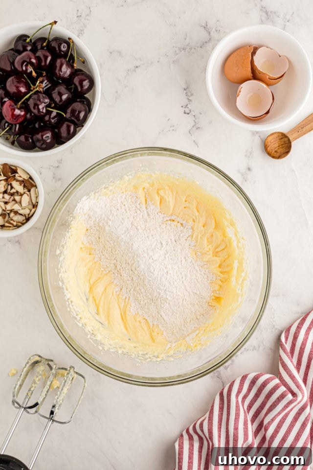 An overhead shot showing the dry flour mixture being gradually added to the wet, creamy butter and sugar mixture in a glass bowl, indicating the start of combining the cake batter components.