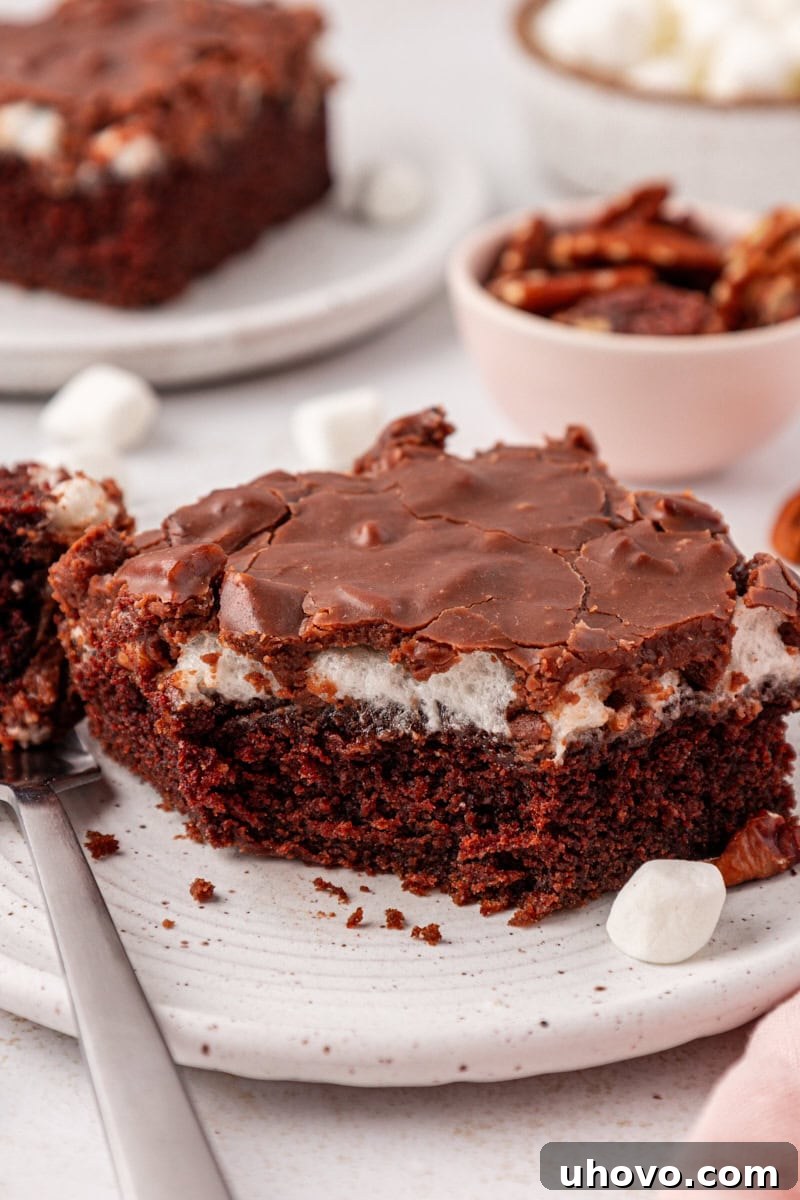 A perfectly sliced piece of Mississippi mud cake on a pristine white plate, with a fork poised for a bite. In the background, more of the glorious cake, sprinkled with pecans and softened marshmallows, awaits.