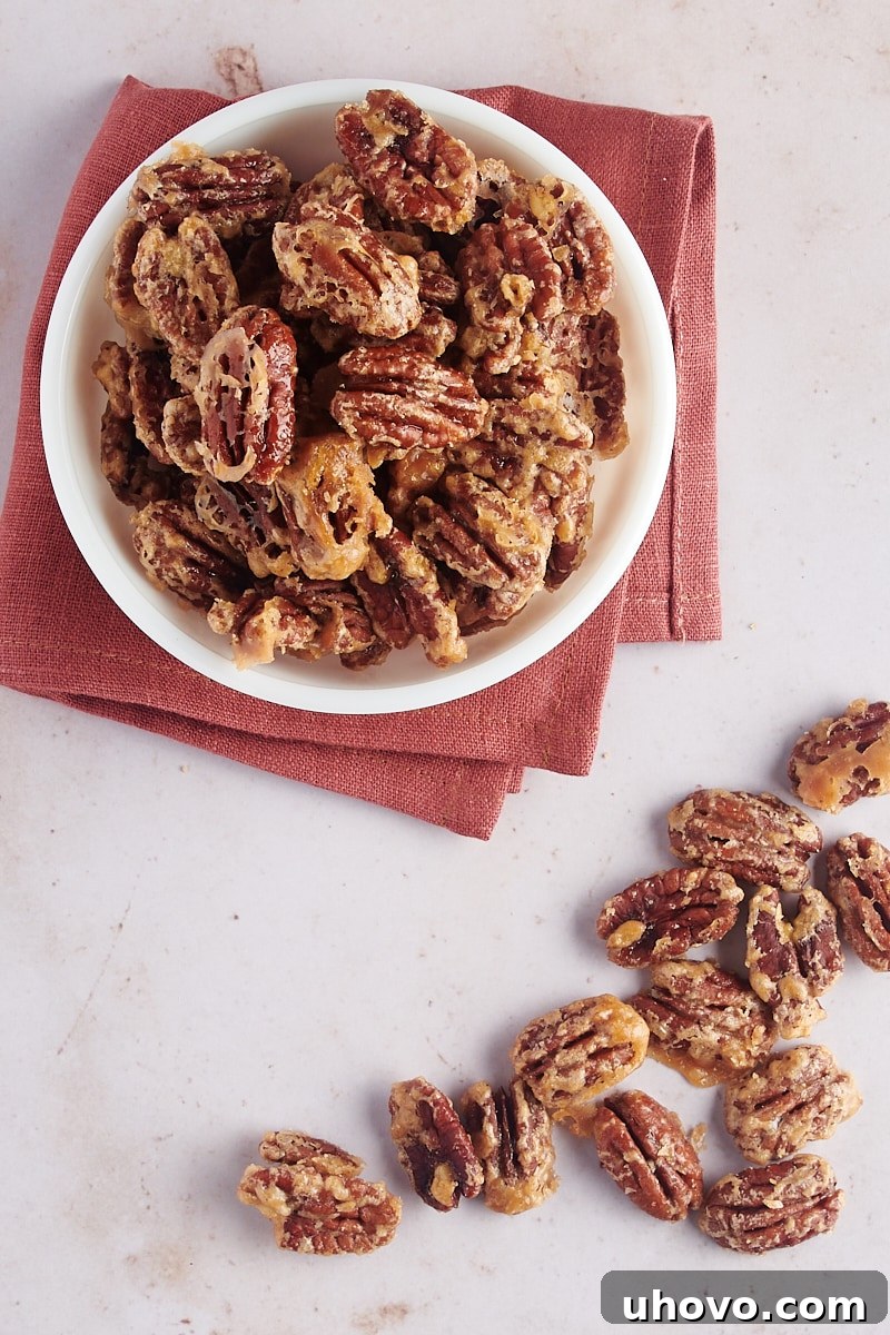 An overhead shot of glazed pecans in a white bowl, with raw pecans scattered beside it on a wooden surface.