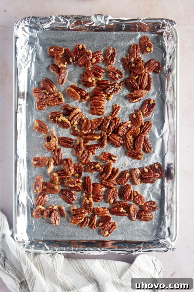 Glazed pecans spread evenly on a foil-lined baking sheet, glistening and ready to be toasted in the oven.