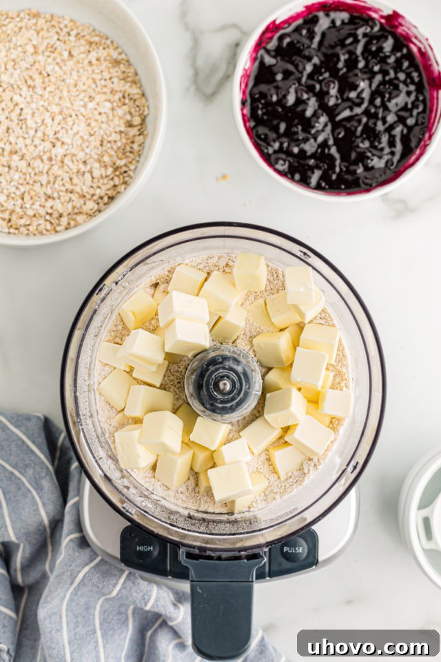 Overhead view of crust mixture with butter cubes in food processor and blueberry jam in bowl