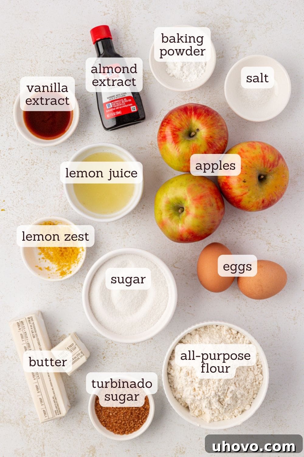 Overhead view of key ingredients for German Apple Cake laid out on a rustic wooden surface, including fresh apples, softened unsalted butter, granulated sugar, all-purpose flour, and small bowls of vanilla and almond extracts, ready for baking.