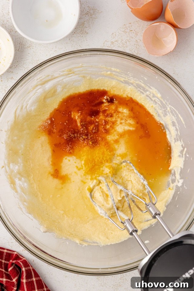 Overhead view of vanilla extract, almond extract, lemon zest, and fresh lemon juice being added to partially mixed cake batter, showing the liquids pooling slightly before being fully incorporated.