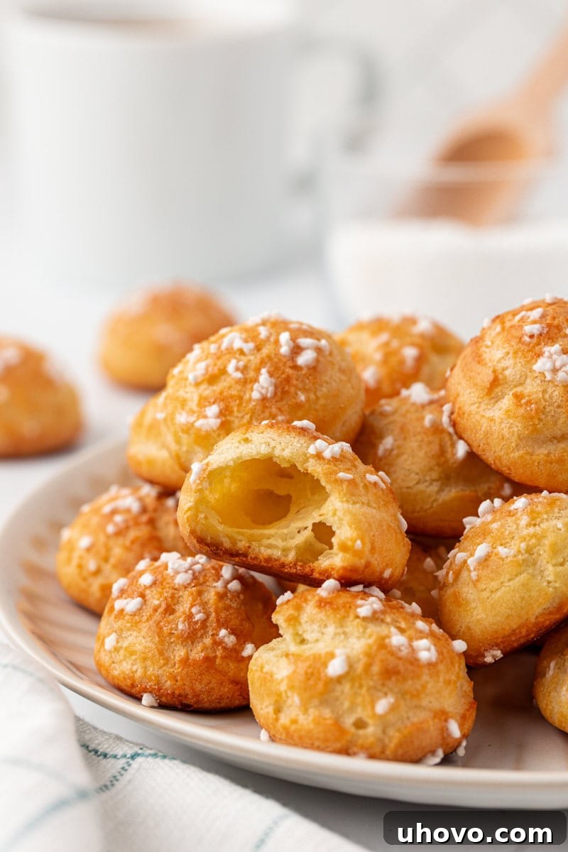 A broken-open chouquette showing the light, airy center with a crisp golden shell, sitting atop more chouquettes arranged on a white plate. This image highlights the perfect texture and inviting appeal of freshly baked chouquettes, ready to be enjoyed.