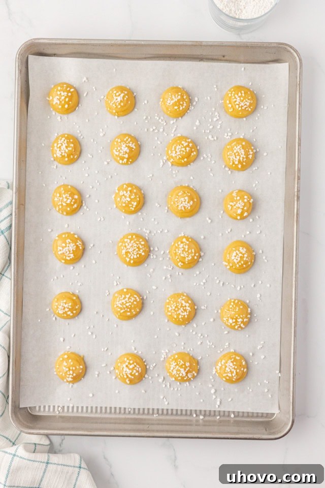 overhead view of piped chouquettes on a parchment-lined baking sheet, brushed lightly with egg wash and generously topped with pearl sugar, glistening and ready for the oven