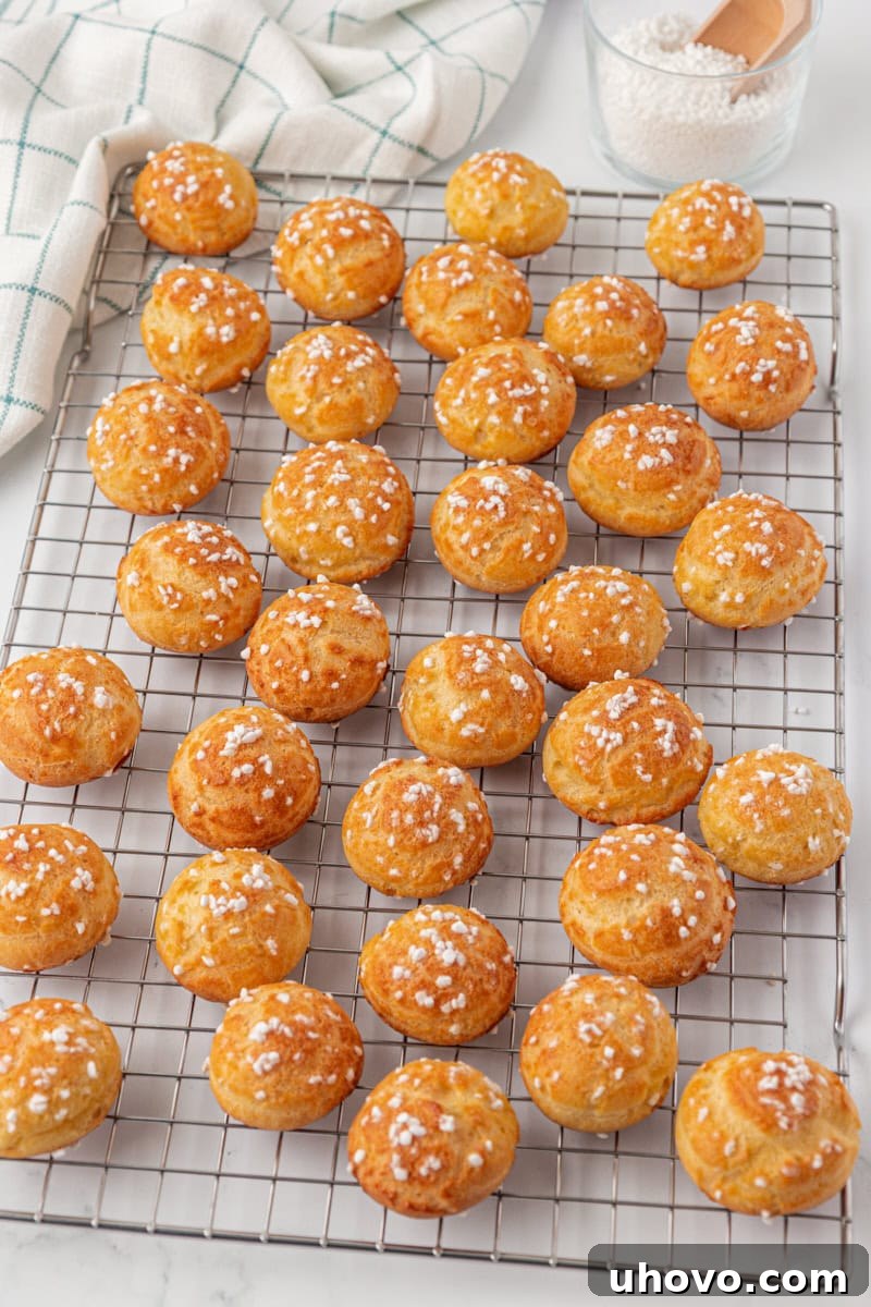 Golden chouquettes cooling on a wire rack after being baked, with sparkling pearl sugar visible on their crisp surfaces, showing their readiness to be enjoyed.