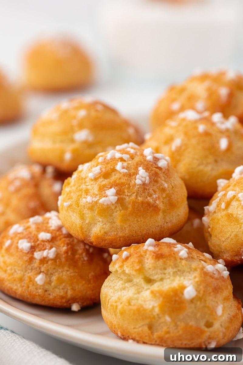 Close-up view of a mound of golden chouquettes, perfectly puffed and covered in glistening pearl sugar, arranged on a pristine white plate, ready to be enjoyed as a delightful French pastry.