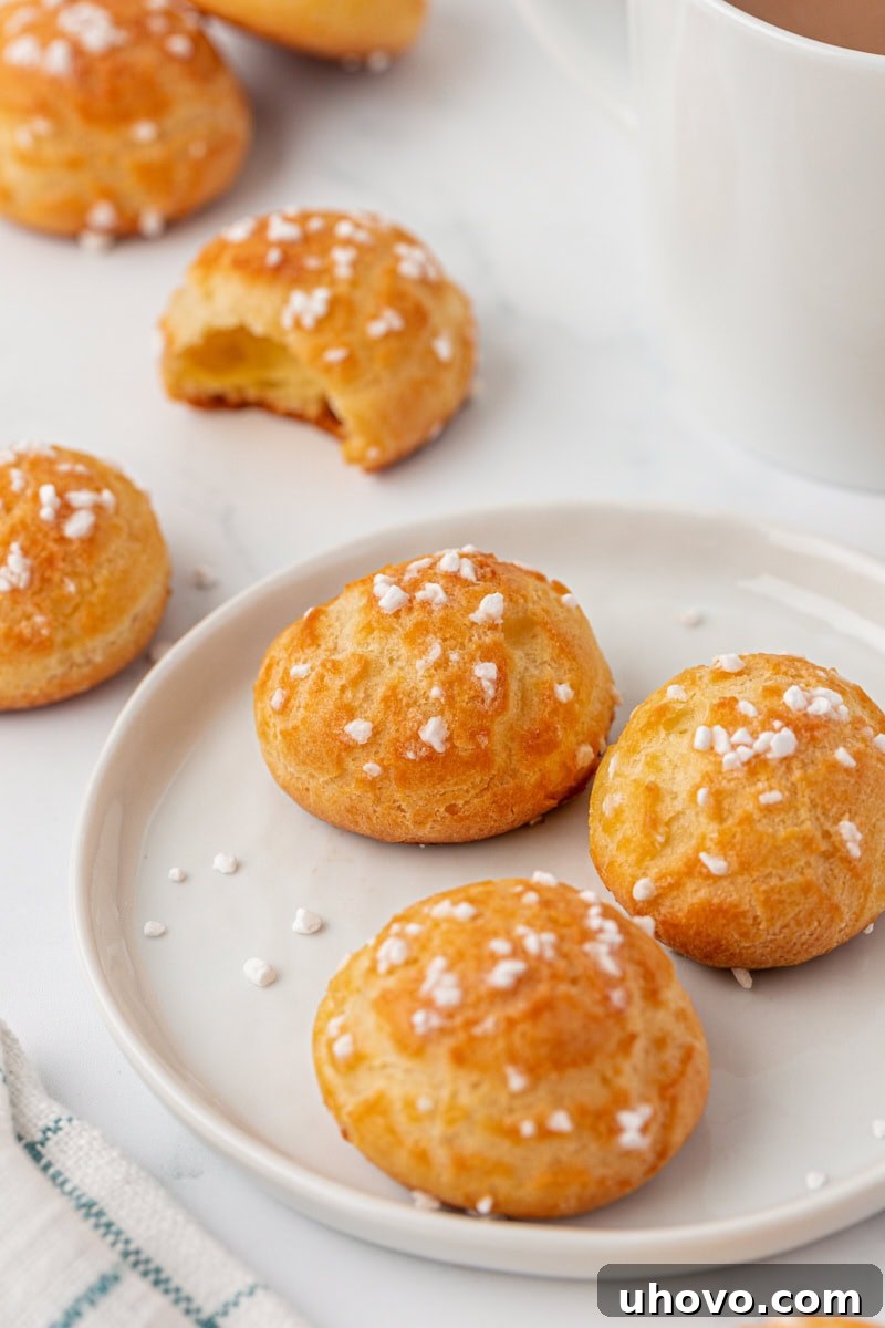 Three golden chouquettes on a white plate, with more pastries and a warm mug of coffee artfully blurred in the background, creating an inviting and cozy scene for a French treat.